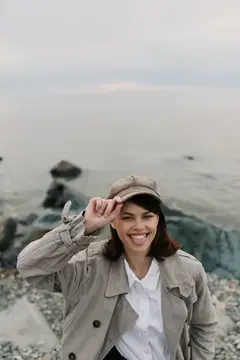 Улыбающаяся темноволосая девушка в кепке и плаще на каменистом берегу