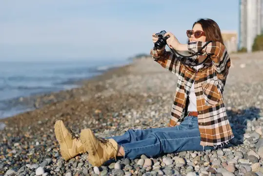 Женщина-фотограф на галечном пляже кадрирует момент у моря с помощью камеры