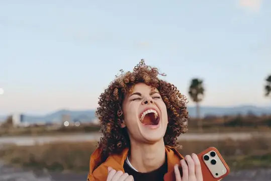 Кудрявая девушка с телефоном громко смеётся на улице