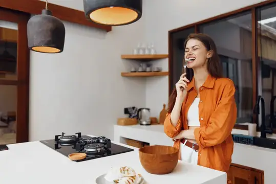 Улыбающаяся девушка в оранжевой рубашке говорит по телефону на кухне