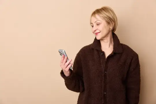Улыбающаяся блондинка в коричневой куртке смотрит в смартфон на бежевом фоне