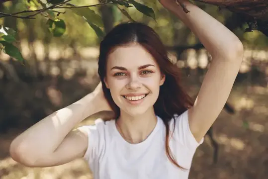 Улыбающаяся темноволосая девушка в белой футболке под деревьями