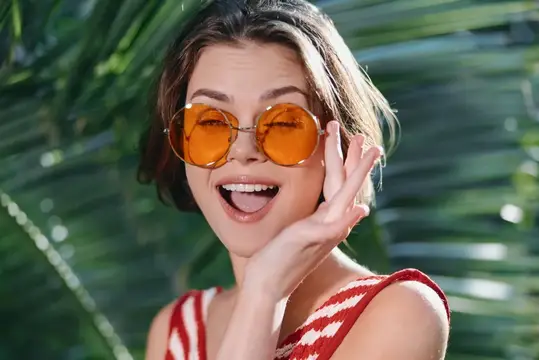 Улыбающаяся девушка в оранжевых круглых очках на фоне пальмовых листьев
