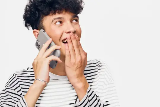 Улыбающийся мужчина в полосатой рубашке держит смартфон, выражая удивление и счастье на фоне однотонного белого фона, запечатлевая радостный момент