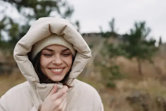 Портрет улыбающейся женщины в теплом пальто с капюшоном на открытом воздухе, расслабленная поза на фоне сельского пейзажа, подчеркивающая уютную ткань, повседневный стиль и безмятежную природную…