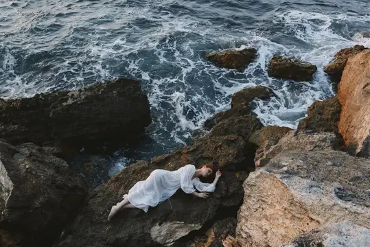 Женщина в белом платье лежит на скалах у бурного моря