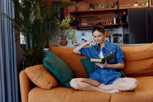 Молодая женщина в джинсовом комбинезоне читает книгу на оранжевом диване