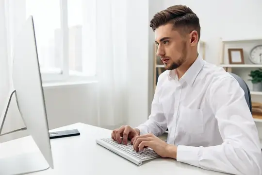 Молодой мужчина в белой рубашке работает за компьютером в светлом офисе