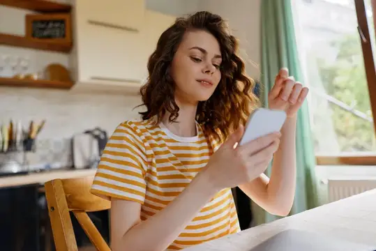 Кудрявая девушка в полосатой футболке листает телефон на кухне
