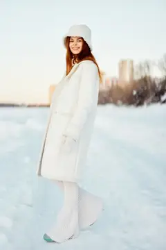 Молодая женщина в белом пальто и шляпе гуляет по зимнему пейзажу, вдыхая свежий воздух
