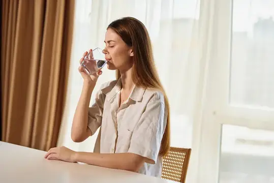 Молодая женщина с длинными волосами пьёт воду из стакана у окна