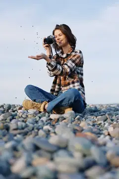 Молодая женщина с фотокамерой сидит на каменистом пляже и подбрасывает камешки