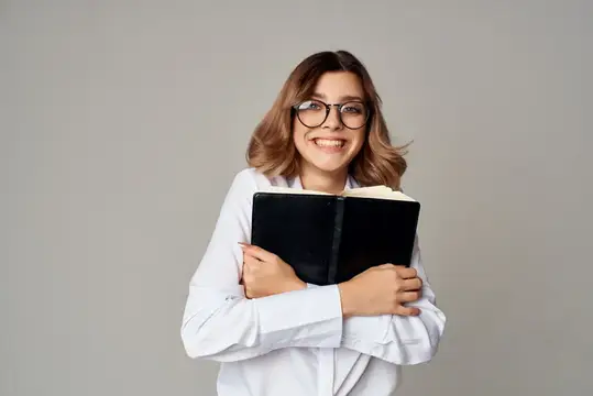 Улыбчивая девушка в очках обнимает чёрный блокнот на сером фоне