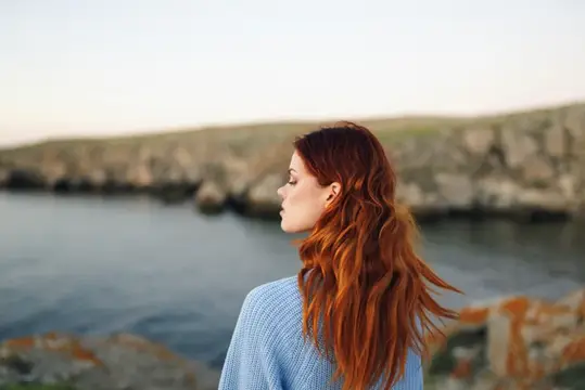 Рыжеволосая девушка в голубом свитере смотрит на море