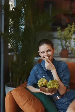 Улыбающаяся девушка в джинсовой куртке ест виноград на диване