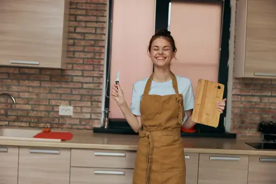 Улыбающаяся девушка в фартуке с ножом и разделочной доской на кухне