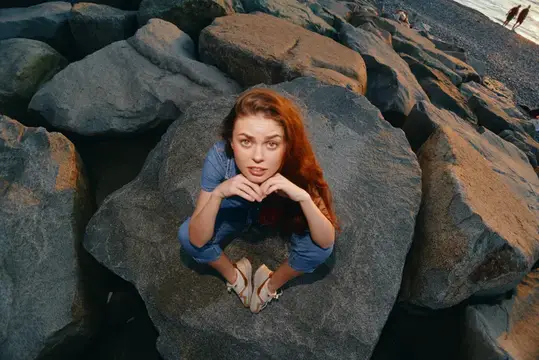 Рыжеволосая девушка сидит на большом камне у моря