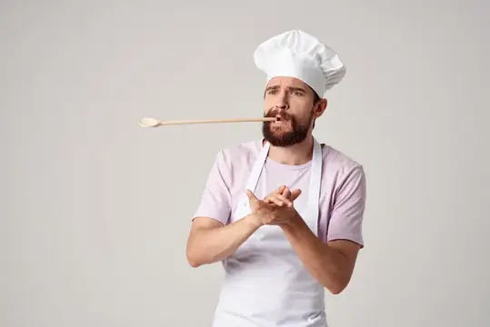 Бородатый повар с деревянной ложкой в зубах на сером фоне