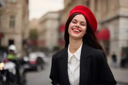 Портрет модной женщины, улыбающейся с зубами, стоящей на улице на фоне города в стильной одежде с красными губами и красным беретом