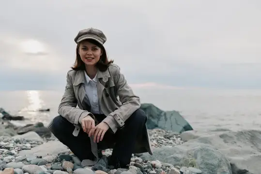 Улыбающаяся девушка в кепке и плаще сидит на камнях у моря