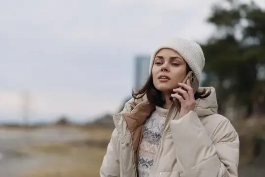 Молодая женщина в пуховике и шапке говорит по телефону на улице