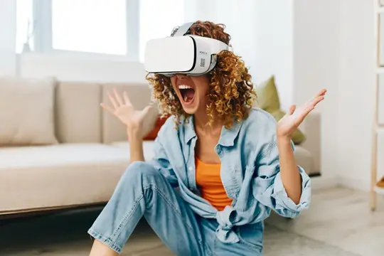 Улыбающаяся женщина наслаждается игрой в виртуальной реальности дома, сидя на диване в VR-очках и с контроллером в руках
