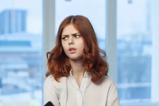 Рыжеволосая девушка в белой рубашке хмурится у окна офиса