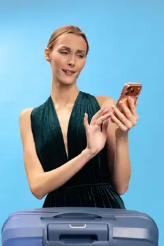 Улыбающаяся светловолосая женщина с телефоном у синего чемодана