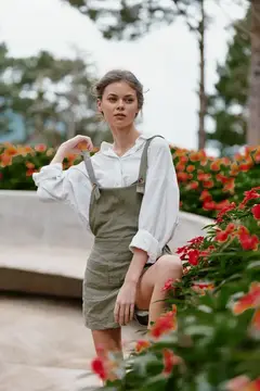 Молодая женщина в оливковом сарафане позирует среди цветущих клумб