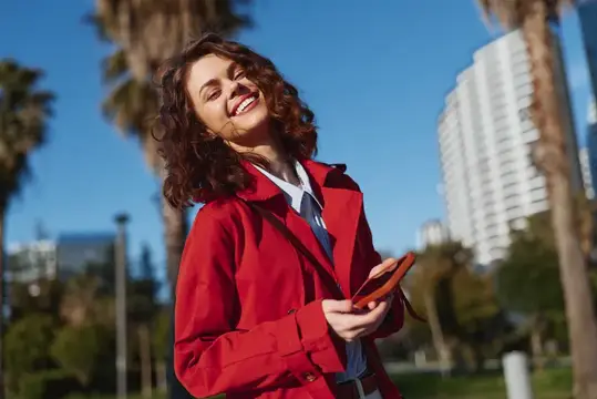 Улыбающаяся кудрявая девушка в красном плаще с телефоном на улице
