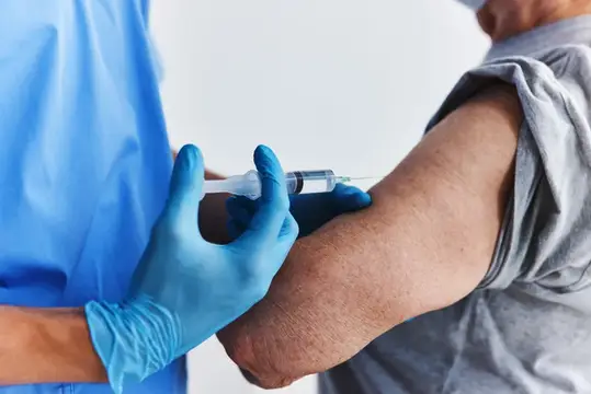 Медик в синих перчатках делает укол в плечо пациенту