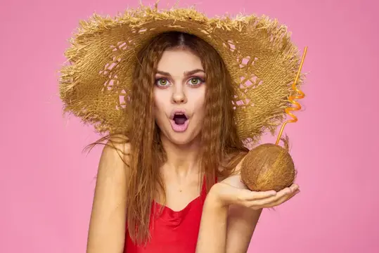 Удивлённая девушка в соломенной шляпе с коктейлем в кокосе на розовом фоне