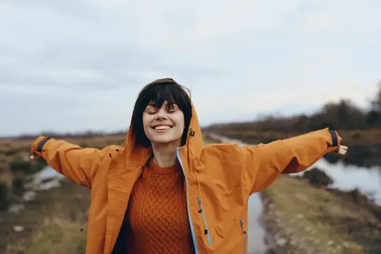 Женщина на открытом воздухе в стиле лайфстайл в оранжевой куртке с капюшоном: улыбается, раскинув руки у воды, осенний пейзаж с облачным небом и рекой, естественный непринужденный портрет в теплом…