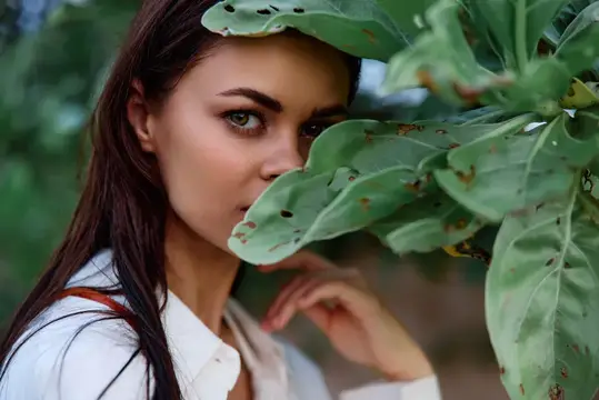 Темноволосая девушка с зелеными глазами выглядывает из-за большого листа