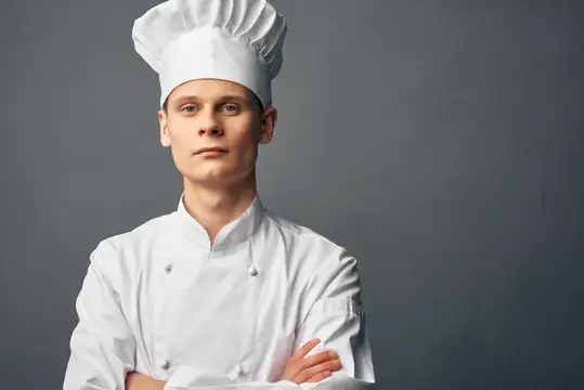 Молодой повар в белом колпаке и кителе на сером фоне