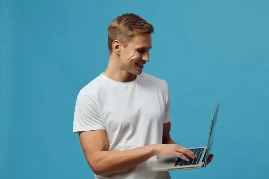 Улыбающийся парень в белой футболке работает за ноутбуком на синем фоне
