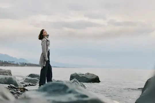 Молодая женщина в плаще стоит на камнях у моря под облачным небом