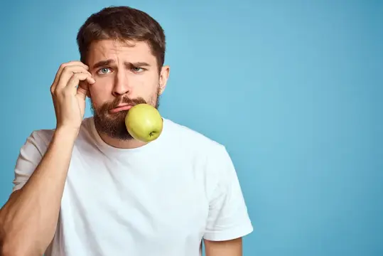 Задумчивый бородатый мужчина с зелёным яблоком у губ на голубом фоне