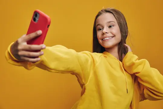 Улыбчивая девочка в жёлтом худи делает селфи на красный смартфон