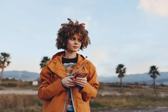 Кудрявая девушка в оранжевой куртке с телефоном на фоне пальм