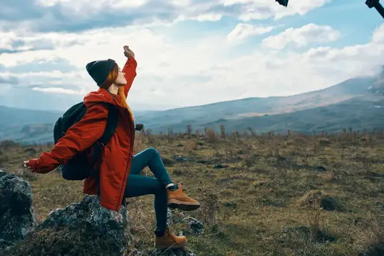 Женщина-путешественница в куртке сидит на большом камне и смотрит на горный пейзаж