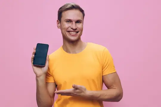 Улыбающийся мужчина в жёлтой футболке показывает смартфон на розовом фоне