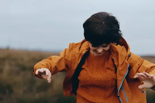 Улыбающаяся девушка в оранжевой куртке танцует в поле
