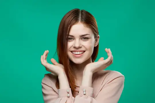 На зелёном фоне симпатичная женщина в розовой рубашке держит руки у лица