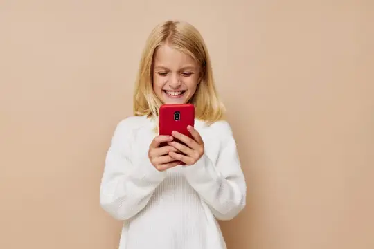 Светловолосая девочка смеётся, глядя в красный смартфон