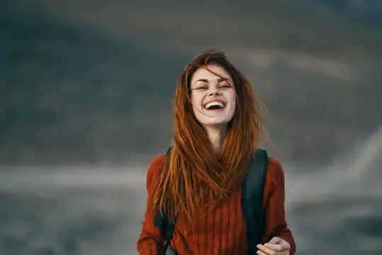 Рыжеволосая девушка с рюкзаком смеётся на фоне гор