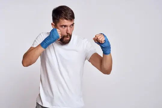 Спортивный мужчина в белой футболке, на руках синие боксёрские бинты, выполняет тренировочные упражнения
