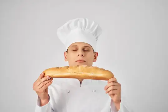 Мальчик в поварском колпаке с закрытыми глазами держит длинный батон