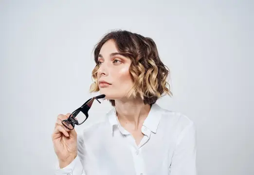 Задумчивая женщина с темными кудрями держит очки на светлом фоне