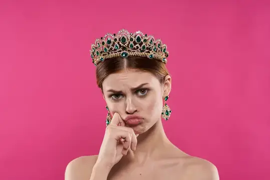 Недовольная девушка в короне с изумрудами на розовом фоне